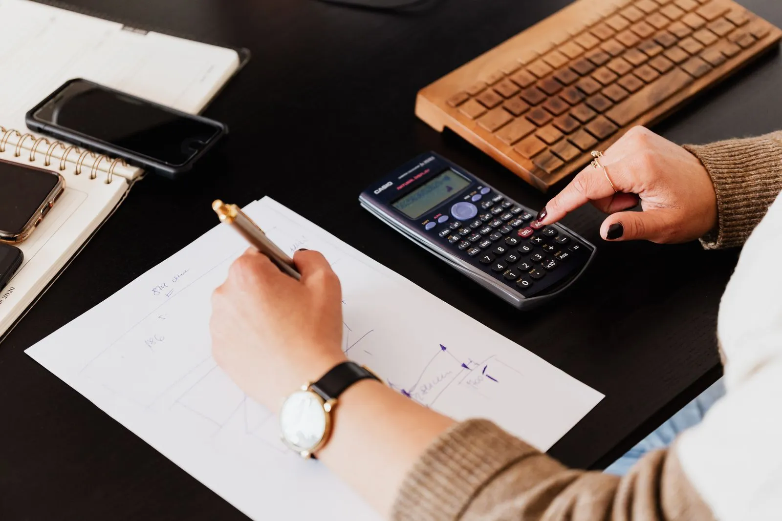 A bookkeeper writing on paper while using a calculator at a desk, illustrative stock photo.